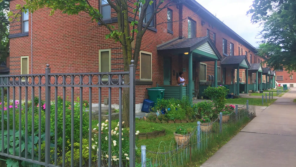 Row of brick townhouses with small front porches, fenced gardens, and a tree-lined sidewalk in a residential neighborhood.