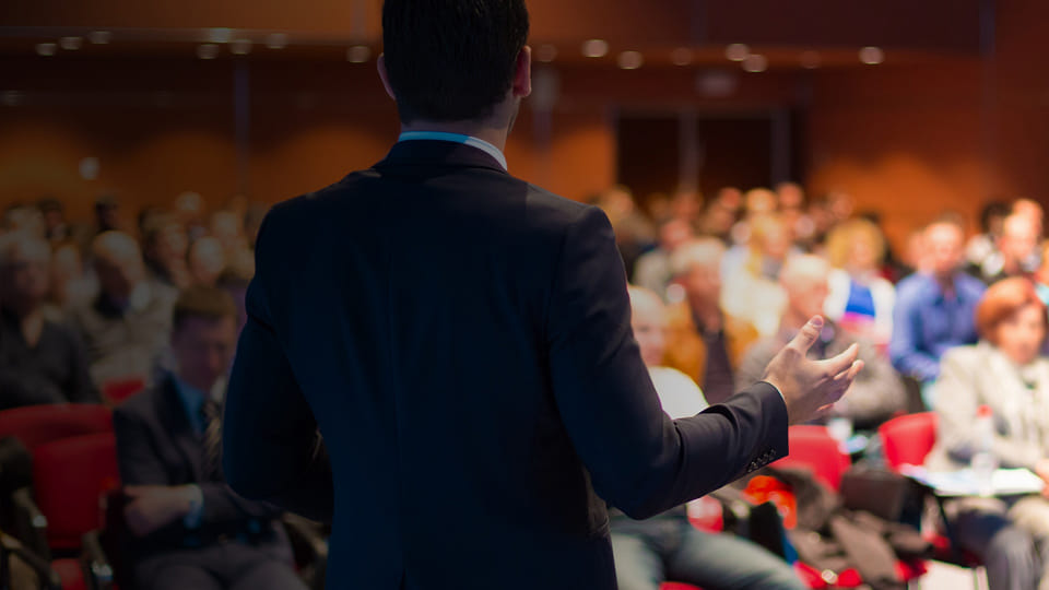 Business professional speaking at a conference in front of a seated audience.